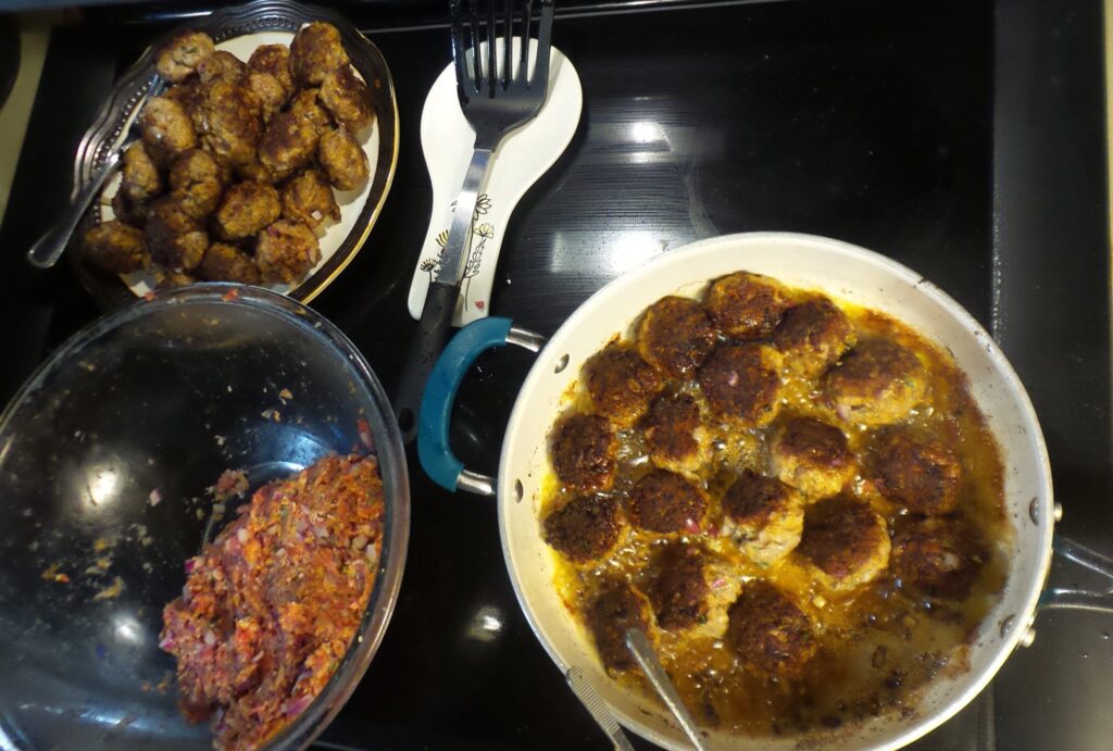 Italian meatballs frying in a pan next to a bowl of ingredients and a plate of cooked meatballs